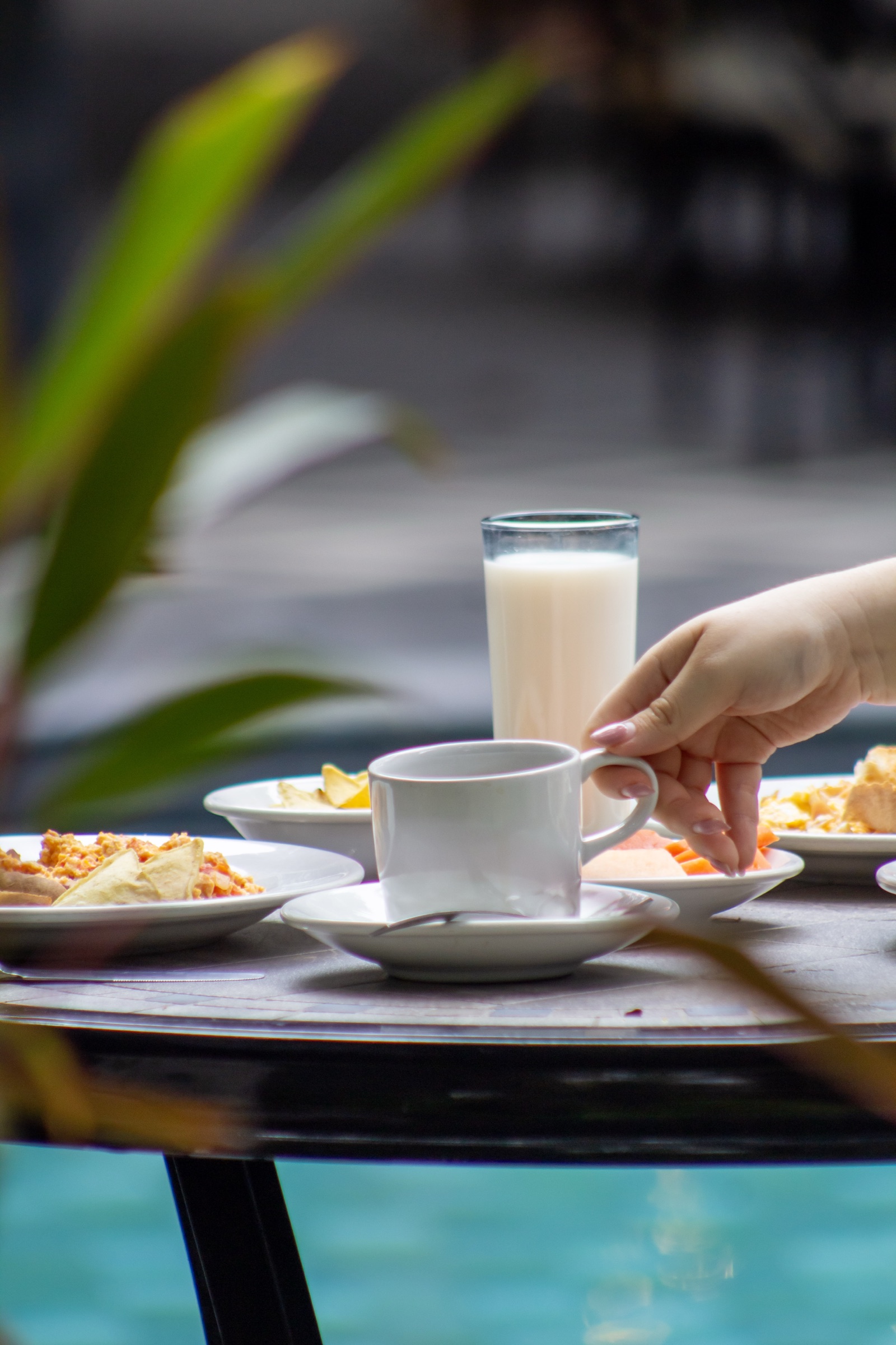 Fotografía de producto de un desayuno: mano tomando una taza junto a un vaso de leche y platos, con una alberca al fondo